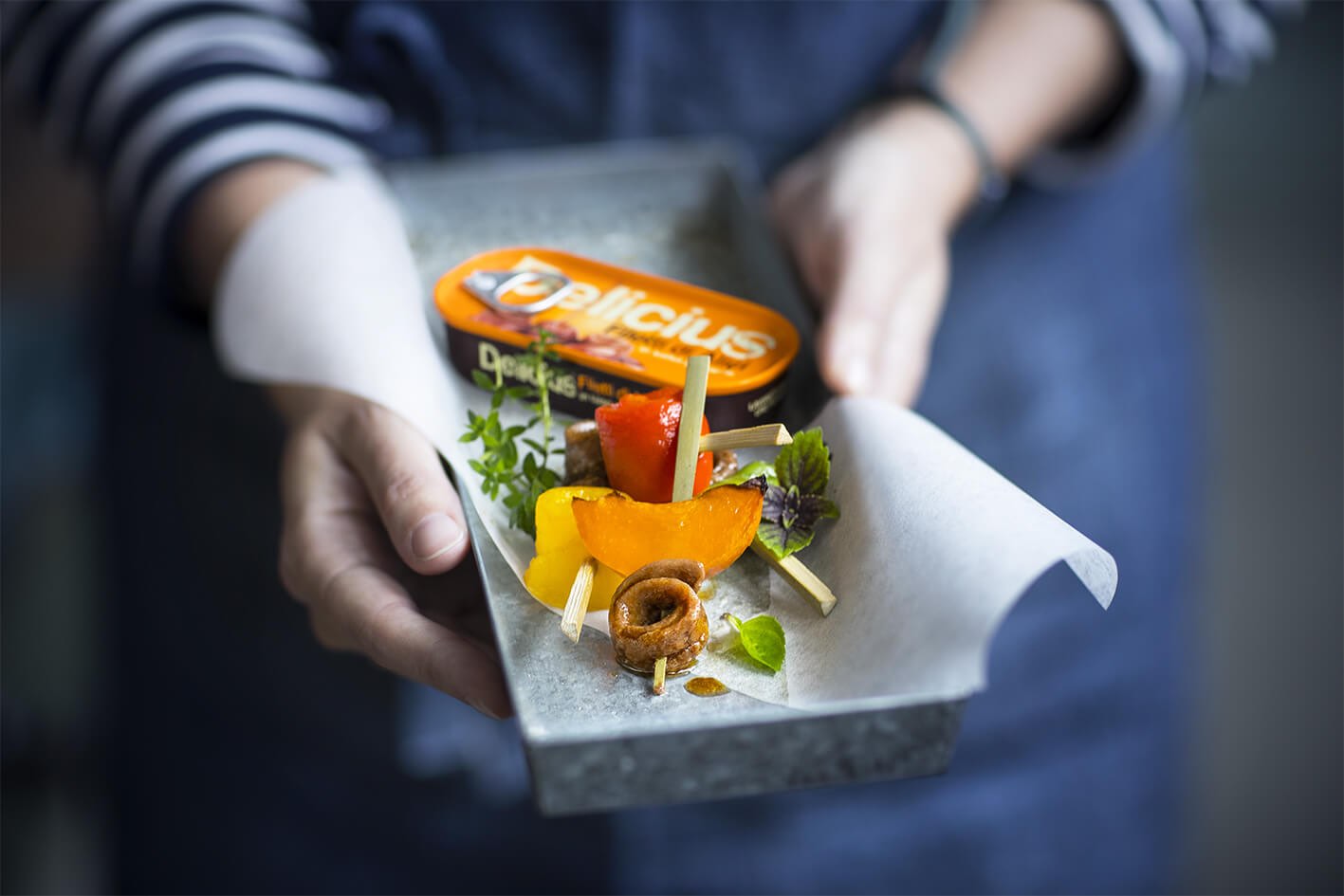 Person holding a small dish with appetizers and a 'Delicius' Anchovies in Spicy Sauce can with label.